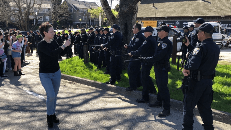 A line of dozens of riot police square off against student protesters at UCSC. Organizer Yulia Gilich stands between the students and the police, recording the police with her cell phone.