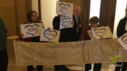 Several community members are holding up individual signs each featuring the name of a person who had been killed by the police. The community members are standing behind a long scroll of paper unrolled horizontally that lists hundreds of names of people killed by police.