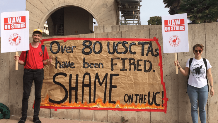 Two striking UCSC graduate students stand on either side of a homemade sign which reads "Over 80 USCS TAs have been FIRED. SHAME on the UC!" in black lettering on a brown field with a red border. Each TA holds a sign reading "UAW on strike!"