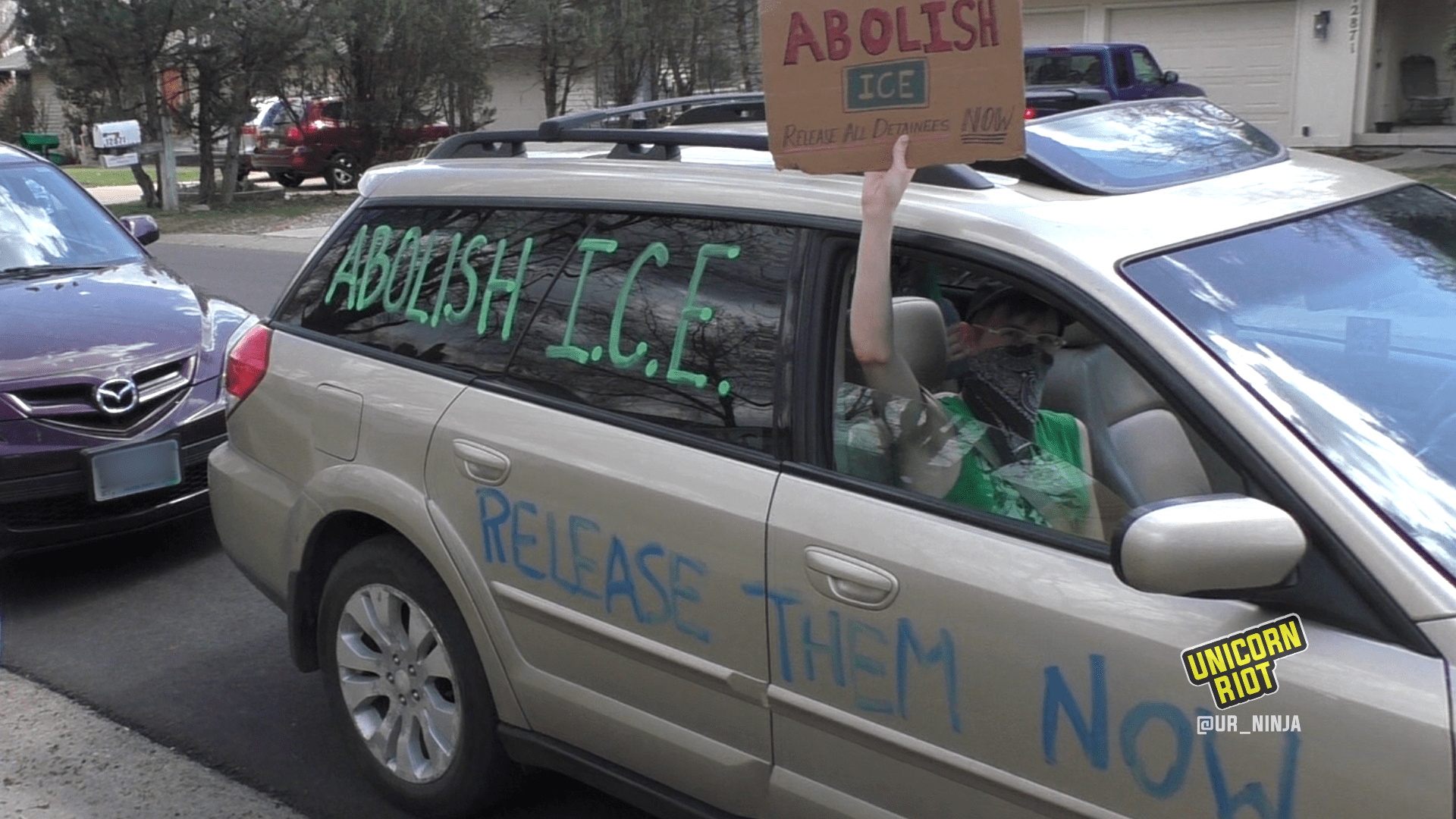 Image description: a gold minivan is painted with the words "Abolish I.C.E." and "Free Them Now" on the windows and sides of the vehicle. A protester's arm holds a sign aloft outside the passenger window; the sign reads "Abolish ICE"