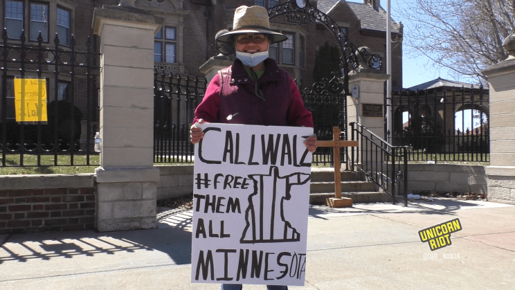 A woman smiles while holding a sign that reads, Call Walz, 'Free Them All Minnesota' with an accompanying drawing of the outline of the state of Minnesota with prison bars. She is wearing a broad-rimmed hat in the sunshine, as well as a medical face mask, which is pulled down to show her smile. She is standing in front of the Minnesota Governor's residence. In the background behind her is a wooden cross, adorned with a crucifix and the initialism INRI, placed at the entrance to the governor's residence.