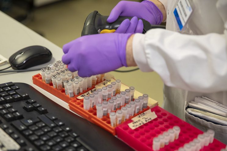 Image of a clinical lab worker scanning barcodes on more than 70 coronavirus testing samples. The worker is wearing purple nitrile gloves, holding the scanner in their right hand and holding a test sample in their left. This photo was taken in Leeds, England in March.