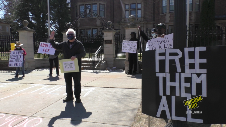 image: a demonstrator with white hair waves his right hand while holding a sign in his left hand that reads 'Free Them All.' He is wearing a face mask made from dark-colored cloth and is standing in front of the governor's residence in Saint Paul. In the foreground is a sign that also reads 'Free Them All' in white text on a black background.