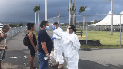 Image: members of the Puerto Rican National Guard wearing masks, eye protection, and white dust suits screen recently-deboarded passengers from an airplane before permitting them to enter Puerto Rico.