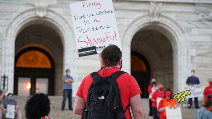 image: A person facing away from the camera holds a sign, "Firing frontline workers in a pandemic is shameful." The person is wearing a red shirt and a black backpack. In the background are protesters standing ~6' apart on the steps of the Capitol building in Saint Paul.