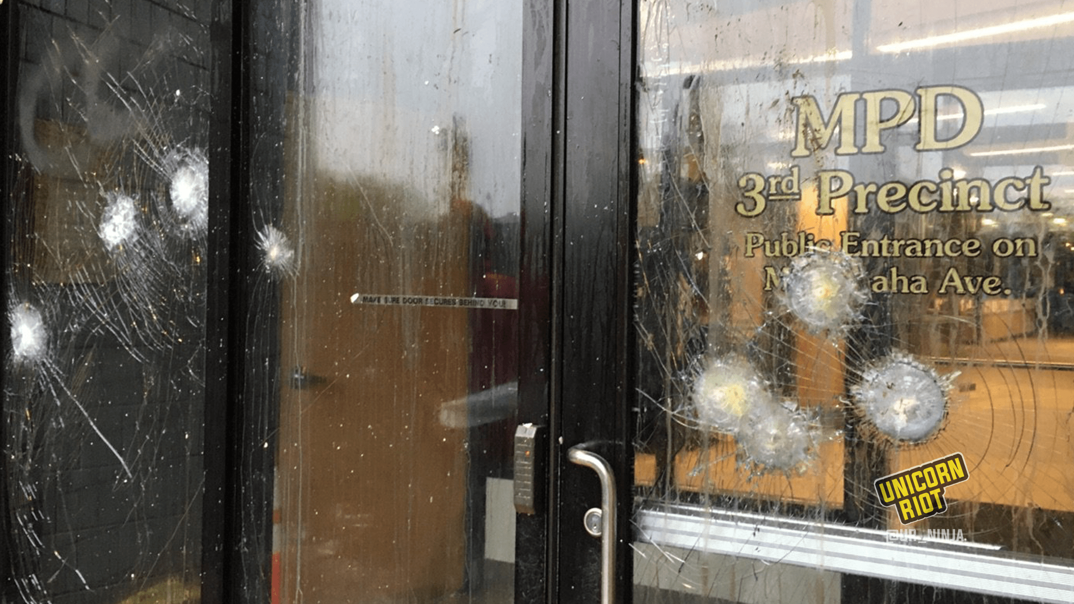 Image: the side entrance to the Minneapolis Police Department uses bullet-proof glass in the windows and door, which are each showing several fractures from having objects thrown at them from protesters. 'MPD 3rd Precinct, Public Entrance on Minnehaha Ave.' is written in gold stencils on the door.