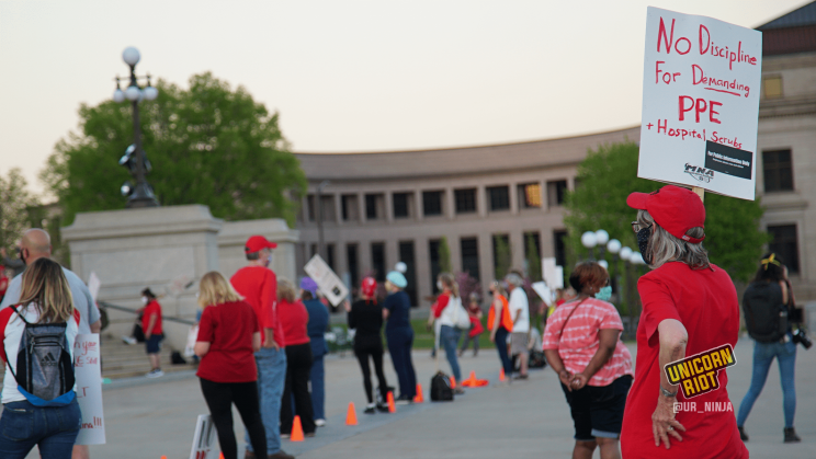 Image: medical professionals and supporters stand in a row, each 6 feet or 2 meters apart from the next, spaced out by orange cones placed on the ground. Every protester is wearing a cloth mask covering their nose and mouth, as this photo was taken during the pandemic of 2020. One person wearing a red baseball cap and red mask holds a sign, "No Discipline For Demanding PPE + Scrubs" with the Minnesota Nurses Association (MNA) logo on the bottom.