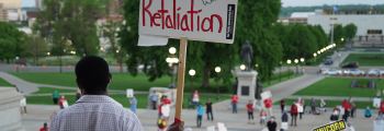 image: a protester stands in front of the entrance to the Minnesota state capitol facing the crowd of protesters, who are spaced out physically-distanced from one another by small orange cones. The protester is holding a sign, "NO retaliation, #BringBackCliffAndMonica, Protect Frontline Workers"