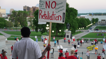 image: a protester stands in front of the entrance to the Minnesota state capitol facing the crowd of protesters, who are spaced out physically-distanced from one another by small orange cones. The protester is holding a sign, "NO retaliation, #BringBackCliffAndMonica, Protect Frontline Workers"