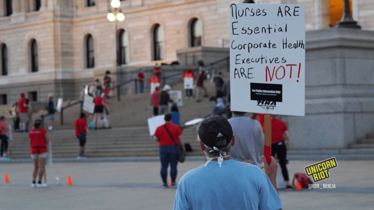 image: a protester stands facing away from the camera, holding a sign: Nurses are essential, corporate executives ARE NOT." The person is wearing a dark blue baseball cap and a light blue t-shirt.