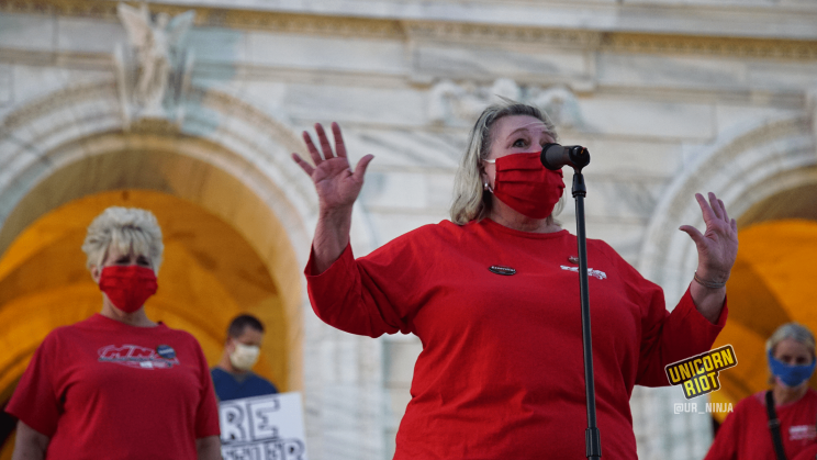 image: National Nurses Association co-president Jean Ross stands in front of a microphone wearing a red long-sleeved shirt and a red mask. She is speaking and has her arms raised at 90 degrees with her fingers splayed slightly facing the crowd.