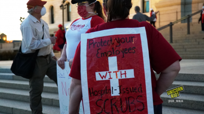 image: a protester stands facin away from the camera. They are wearing a red shirt, over which is a red-and-white sign. The border is white, with a white cross in the center of a red field. There is black and red text overlaid, "Protect your employees with Hospital-issued scrubs"