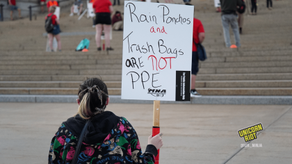 A protester at the informational picket is seated on the ground holding a sign "Rain Ponchos and Trash Bags are NOT PPE." The protester is wearing a black hooded sweatshirt with a pattern of red flowers and green foliage, and has their hair in a ponytail. In the background are protesters standing ~6' apart on the steps of the Capitol building in Saint Paul.