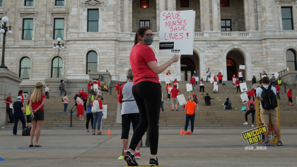A protester at the informational picket turns around and looks backwards, holding a sign "Save Nurses Save Lives." She is wearing glasses, a gray patterned mask a red shirt and black pants. In the background are protesters standing ~6' apart on the steps of the Capitol building in Saint Paul.