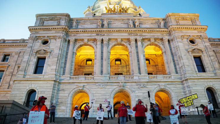 image: protesters stand on the steps in front of the entrance to the Minnesota Capitol building. The sun is setting and as the sky grew darker the lights on the grounds turned on, including the building itself which is backlit with an orange/yellow glow behind the people.