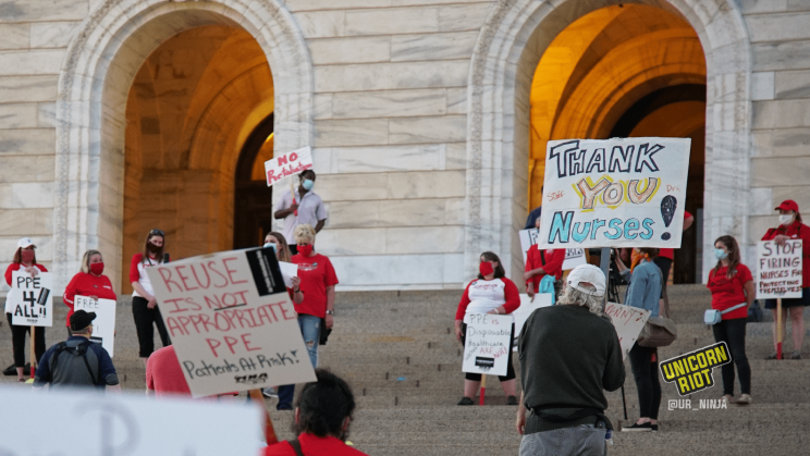 Image: two signs held by two different protesters (both of whom are facing away from the camera): first, in the foreground on the left, "Reuse is NOT Appropriate PPE;" secondly, behind it on the right, "Thank You Nurses!"