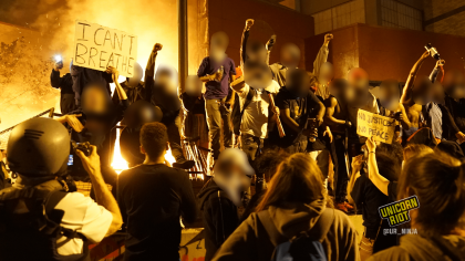 image: dozens of protesters raise their fists as first burn behind them on the grounds of the 3rd precinct. Photo taken at midnight on May 29, 2020