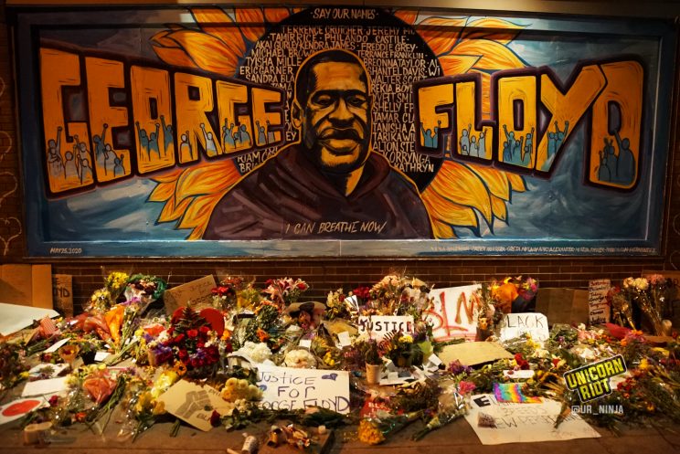 image: photo of large George Floyd mural shows a portrait of Floyd, with his name George on the left and Floyd on the right of his image. A huge sunflower is behind all of this. Painted in the center of the sunflower are many other names lost to police violence.