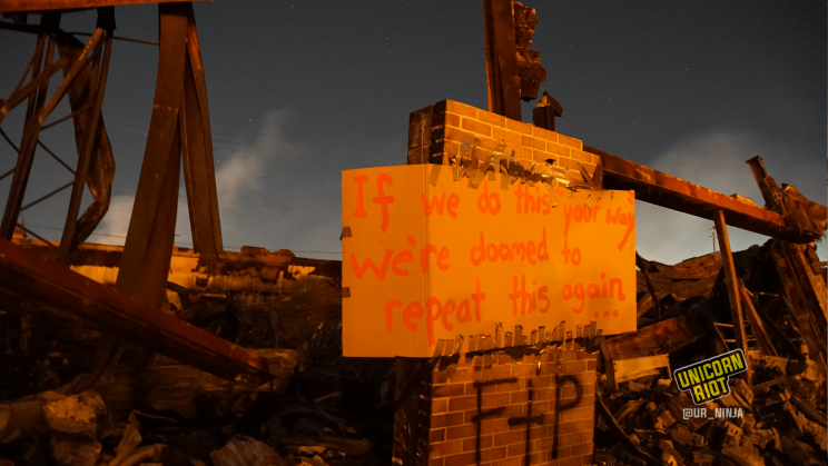 image: a protest sign has been affixed to a tower of bricks still standing amidst the burned rubble of the AutoZone: "If we do this 'your way' we're doomed to repeat this again..." Behind the remains of the building, stars are visible in the night sky.