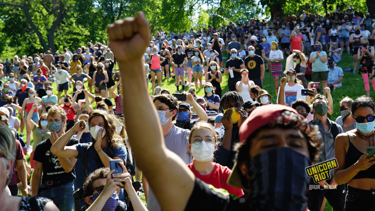 image: the hill of protesters reacts positively to the notion of defunding & disbanding the Minneapolis police department. One person in the foreground has their right arm raised over their head making a closed fist.