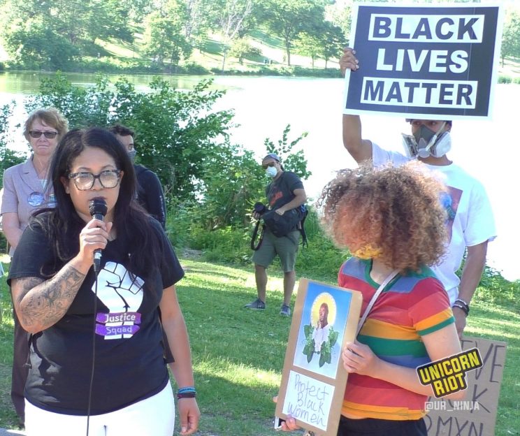 Ashley Quinones speaks while woman holds sign with Oluwatoyin Salau's image and Protect Black Women in words