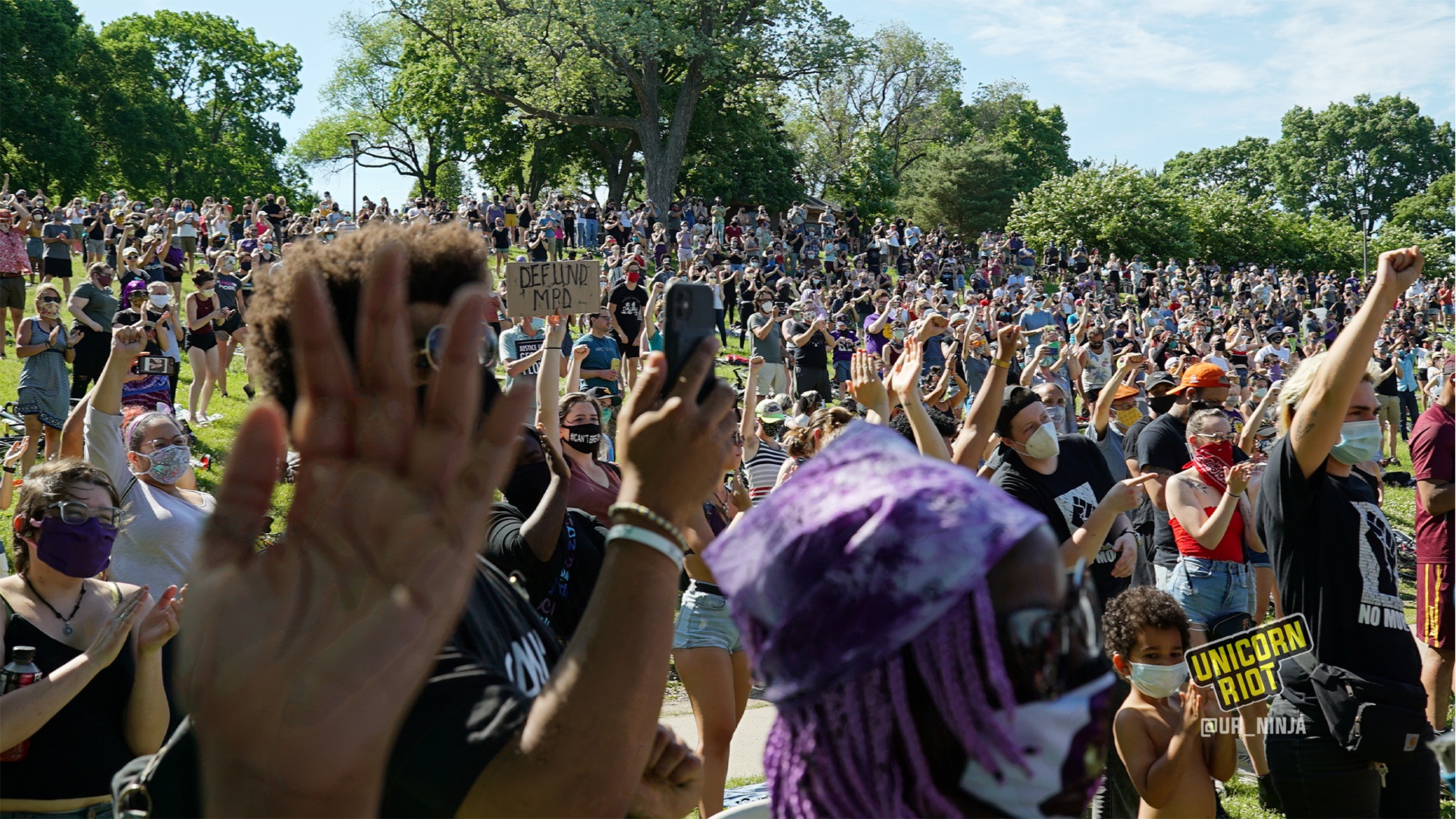 image: the hill of protesters reacts positively to the notion of defunding & disbanding the Minneapolis police department. In the foreground one person's left hand is upraised with their fingers outstretched and their palm towards the stage.