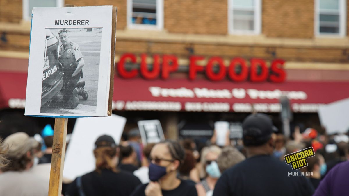 image: hundreds of protesters stand at the intersection of 38th & Chicago in Minneapolis. Everyone is wearing a cloth or paper face mask. In the background is the store front CUP FOODS, in the foreground is a sign with a black-and-white photograph of Derek Chauvin kneeling on the neck of George Floyd in front of the police vehicle; a screenshot from the viral video that shows Floyd's death. The word MURDERER is written above the photograph.