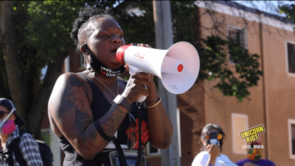 image: a Black woman speaks into a megaphone. She is wearing a fabric mask on her face which is pulled down to allow her speech into the megaphone to be unimpaired. The black fabric mask reads "Guns Down Love Up" -- all the words are white except love is in red. Behind her a protester wearing a mask takes a photo using her smart phone.