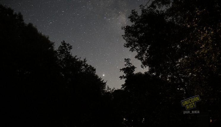 image: ten-second long exposure shot of the night sky facing South, in the northern hemisphere at midsummer. The planet Jupiter is visible with the unaided eye at the bottom of the tree line, below the constellation Sagittarius. The nebulous arm of the Milky Way galaxy is visible to the right and above stretching up to the left away from the planet.
