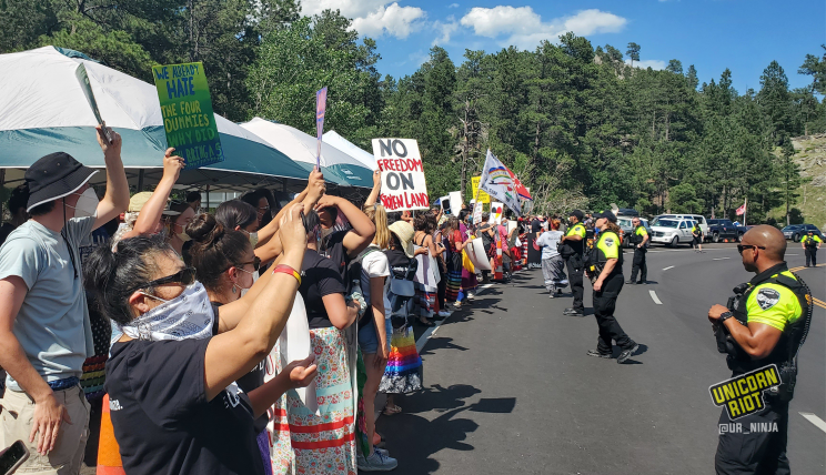 image: Indigenous people and activists gathered on the side of the road leading to Keystone, SD. The anti-Trump, anti-Mount Rushmore demonstrators hold protests signs such as "We already hate the four dummies, why did you bring a 5th?"