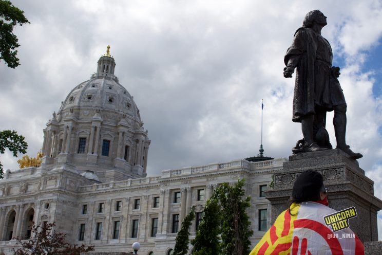 Man with American Indian Movement flag drapped across his back looks at Columbus statue before it got taken down