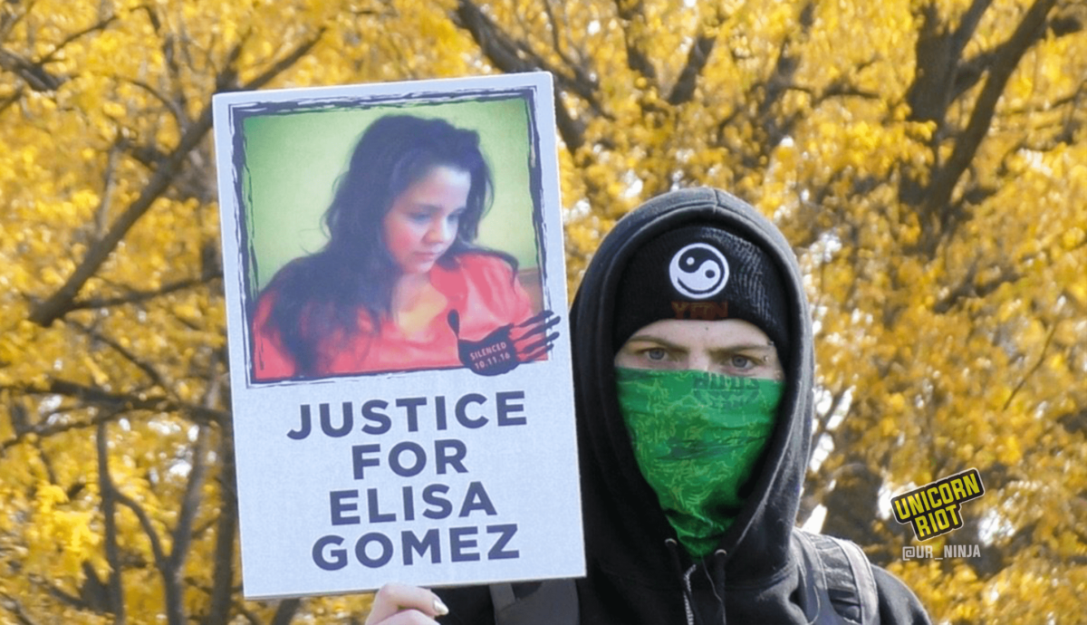 image: Masked protester holds "Justice For Elisa Gomez" protest sign. The sign has a picture of Elisa Gomez with long dark hair and an orange blouse, looking down and to the right. It also has an image of a black handprint with "Silenced 10.11.16" white text overlaid. The protester is wearing a black hooded sweatshirt with the hood up, as well as a black knit cap with a black-and-white Yin Yang symbol, and a green cloth face scarf. They are staring directly into the camera; they have light-colored eyes and white skin, and shiny polish on their nails. Behind them out of focus is a canopy of tree leaves, all turned yellow because of the autumn season.