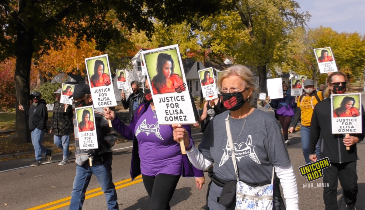 image: Masked protesters hold "Justice For Elisa Gomez" protest signs. Each sign has a picture of Elisa Gomez with long dark hair and an orange blouse, looking down and to the right. It also has an image of a black handprint with "Silenced 10.11.16" white text overlaid. More than a dozen protesters are pictured, many wearing black cloth face masks with a red handprint on the front.