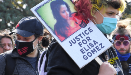 image: Masked protester holds "Justice For Elisa Gomez" protest sign. The sign has a picture of Elisa Gomez with long dark hair and an orange blouse, looking down and to the right. It also has an image of a black handprint with "Silenced 10.11.16" white text overlaid. The protester has on a blue medical facemask, is wearing a black sweatshirt and a purple backpack, and has shoulder-length slightly-curled hair that is dyed yellow and pink. They are looking to the right. Several other masked protesters are behind this person.