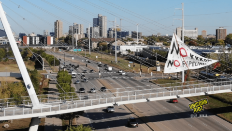 image: a white triangular banner hangs from the cables above the suspension bridge over Hiawatha Avenue in Minneapolis, oriented towards the right. "NO KKKOPS, NO PIPELINES" is written in mostly-black text; in both of the NOs, the letter O is in red. Red "DANGER" caution-type plastic tape is attached at the top point of the triangle and at the point on the right. The city skyline is visible behind the bridge, and there are cars on Highway 55/Hiawatha Avenue below the pedestrian bridge.