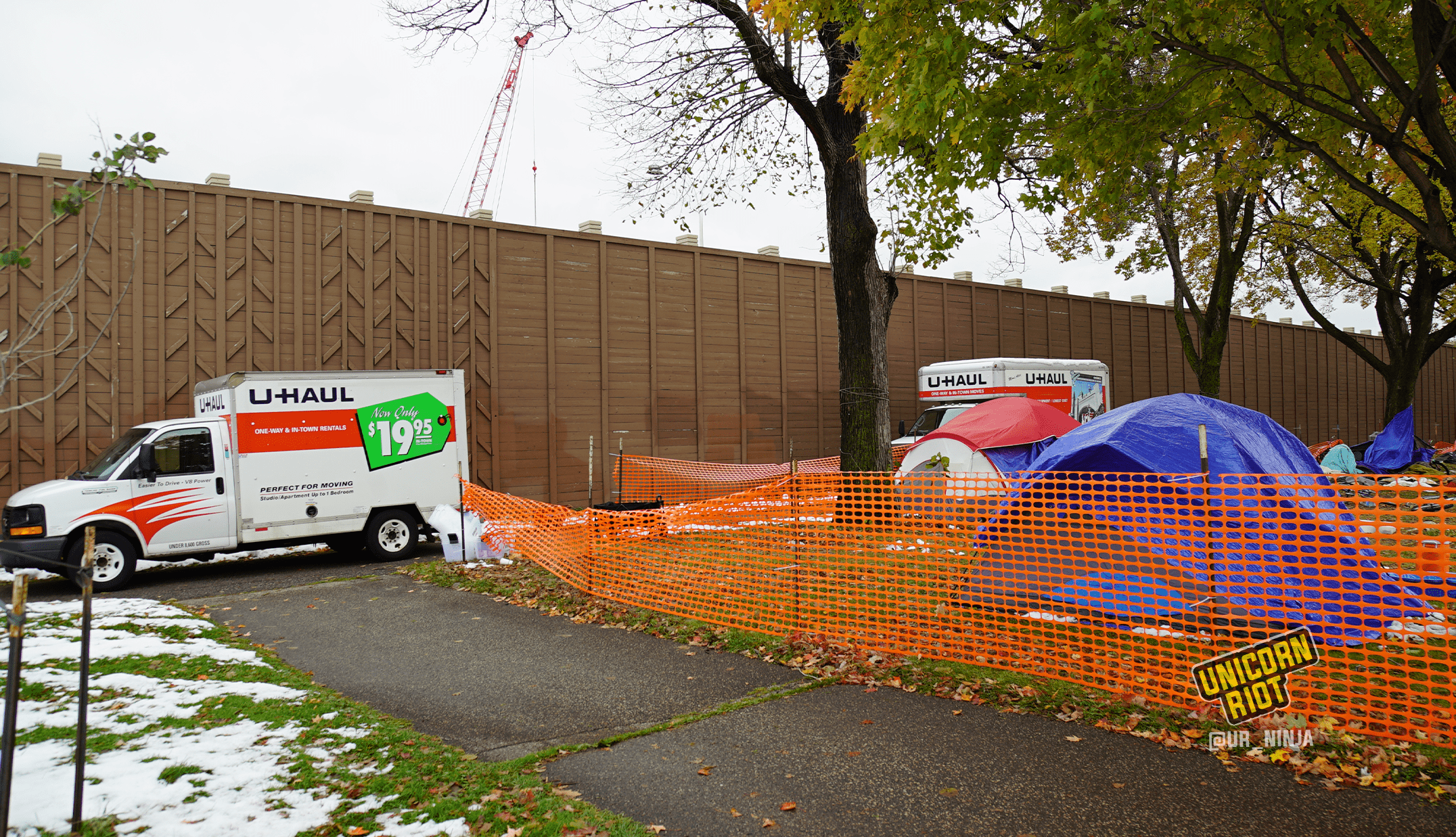 image: the perimeter of a sanctuary tent camp area is surrounded by orange plastic construction fencing; the fence cuts diagonally across the image from bottom right, to the left of the image. On the sidewalk outside of the fencing, two U-Haul trucks are parked; residents' belongings are inside, being moved from MLK Park to another location. A gray tent with a blue tarp is propped up beneath and to the left behind the trunk of a maple tree. To the right of the tree is a waste bin that says City of Minneapolis on its side. Partially-melted snow is visible on the ground.
