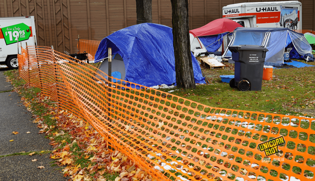 image: the perimeter of a sanctuary tent camp area is surrounded by orange plastic construction fencing; the fence cuts diagonally across the image from bottom right, to the left of the image. On the sidewalk outside of the fencing, two U-Haul trucks are parked; residents' belongings are inside, being moved from MLK Park to another location. A gray tent with a blue tarp is propped up beneath and to the left behind the trunk of a maple tree. To the right of the tree is a waste bin that says Property Of the City of Minneapolis on its side. Other tents are visible in the background.