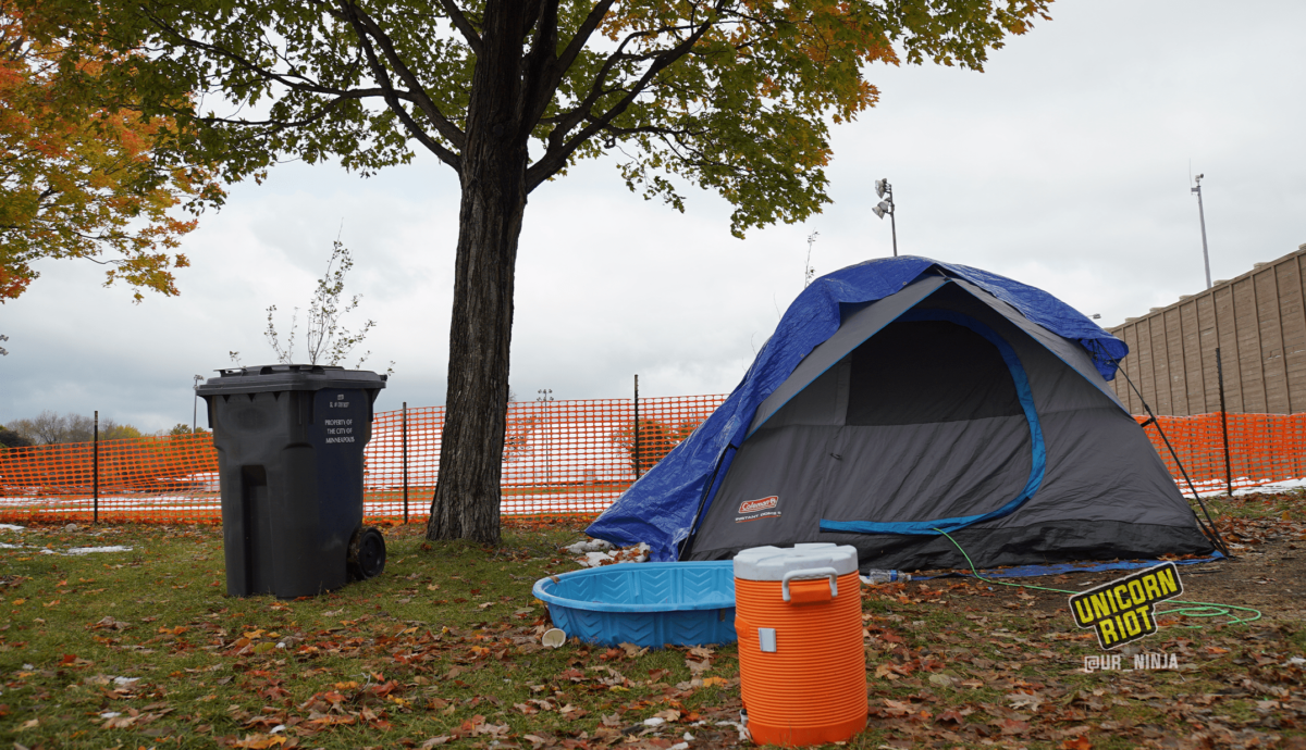 image: a blue and gray tent is propped up beneath and to the right of a maple tree, whose leaves are changing from lime green to yellow, orange, and red tones.  In front of the tent is a children's blue plastic swimming pool, and in front of the pool is an orange water cooler. To the left of the tree is a waste bin that says City of Minneapolis on its side. The entire site is surrounded by orange plastic construction fencing.