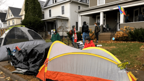 image: single- and double-person tents are positioned on the sidewalk in front of Commissioner Marion Greene's home in South Minneapolis. 5 community members are gathered on her front lawn talking, while a few others are sitting at a table on her front porch.