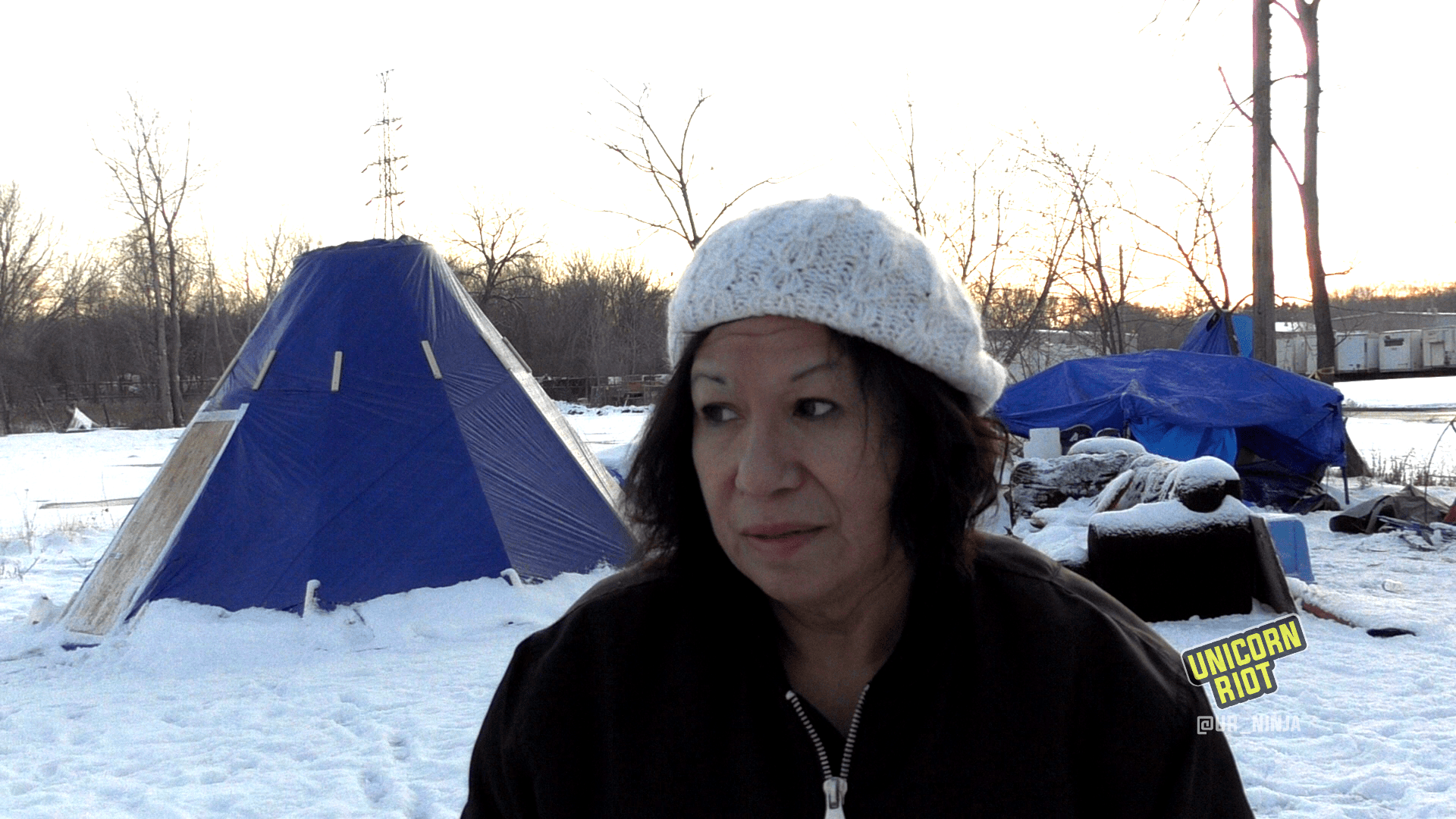 image: Brenda a 60-year-old Native American woman speaks in the center of the frame. She is wearing a white knit hat and a black zip-up jacket. Behind her on the ground is white snow, and to her left in the background is an Indigenous structure known as a Tarpee, a tipi-like structure made with tarps constructed to help elders keep warm while fighting oil pipeline extraction on Native land.