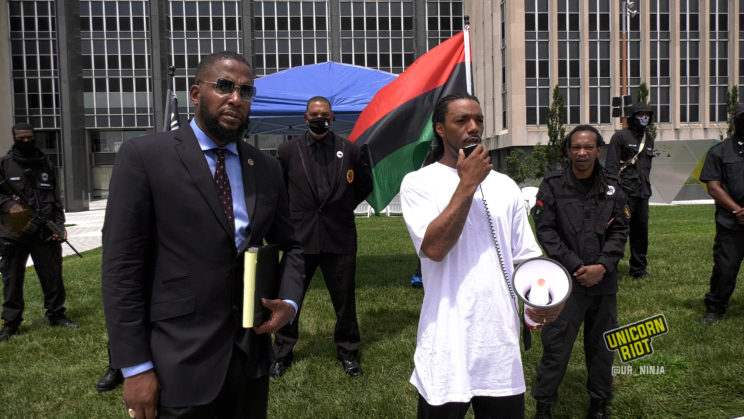 Corey, Dorian Murrell's cousin speaks into megaphone while standing next to family attorney Malik Shabazz and in front of a formation of Black Panther members, some of whom are armed