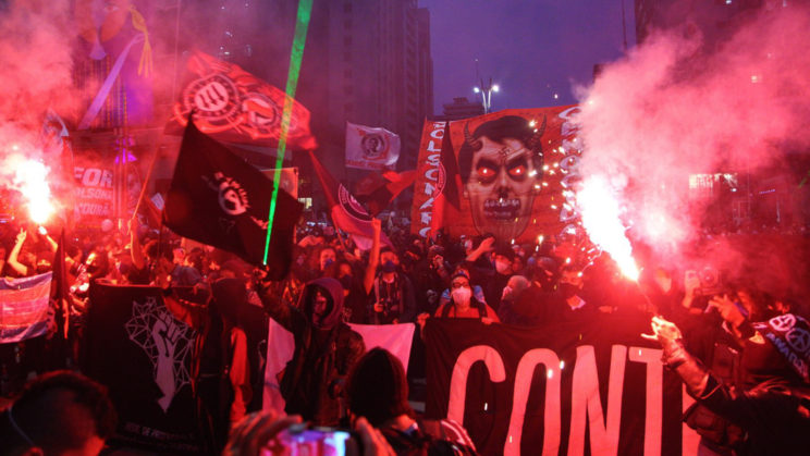large street protest in sao paulo showing flares lit, lazers shining and large signs against brazilian president bolsonaro