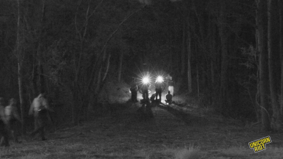 An infrared black-and-white image shows detainees seated in a wooded path in the South River Forest on March 5, 2023 as they are surrounded by police, likely in the process of arresting them. Police flashlights facing the camera create two star-type lighting effects in the photograph. 