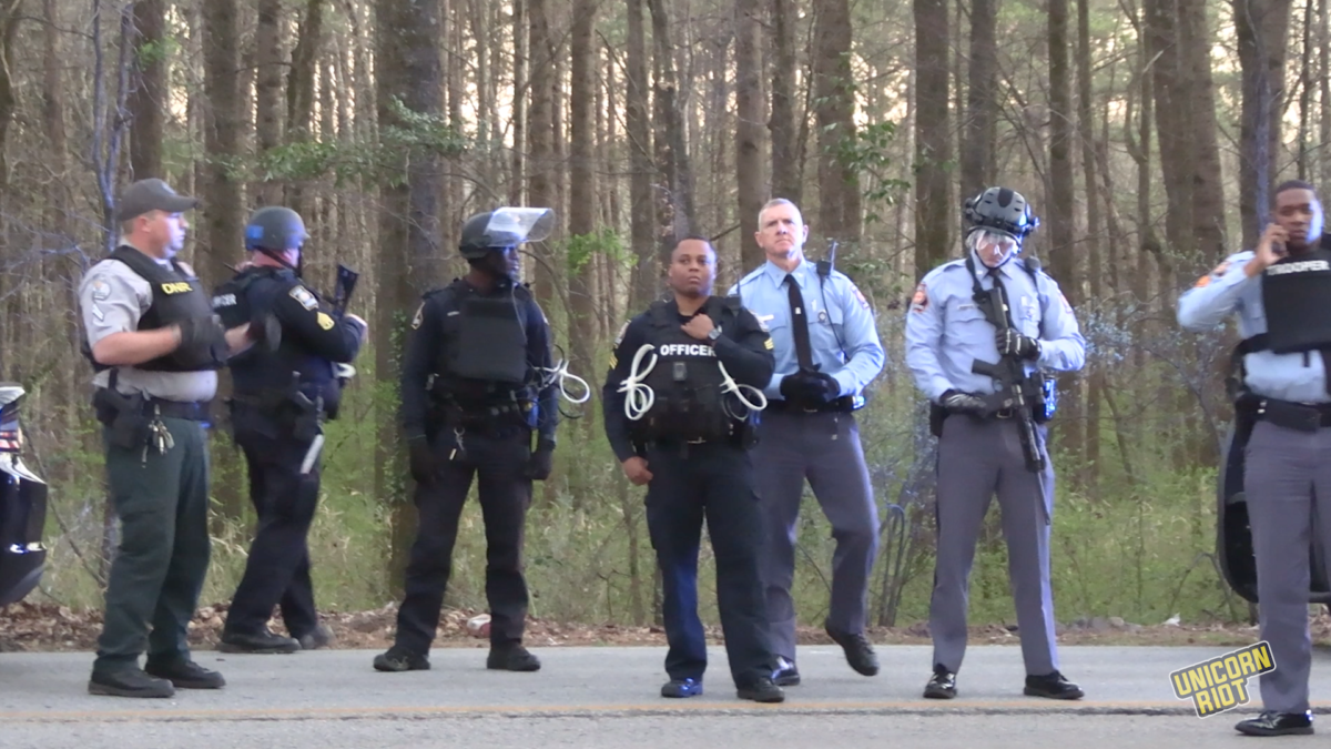 Georgia State Troopers, Department of Natural Resources Game Wardens, DeKalb County Police and Atlanta Police officers stand on a forest road with plastic zip-tie handcuffs attached to their vests. Some have assault rifles. A Georgia State Patrol higher-up with gray hair is seen exercising command while wearing a black tie and gloves, third from the right.