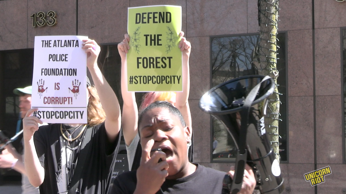 Reverend Keyanna Jones, a Black woman, speaks into a black megaphone as two young white protesters stand behind her holding up signs that say "The Atlanta Police Foundation is Corrupt" and "Defend the Forest." This photo shows these three at a protest outside the downtown Atlanta headquarters of 'Cop City' sponsor corporation Georgia Pacific's offices at 133 Peachtree St. 