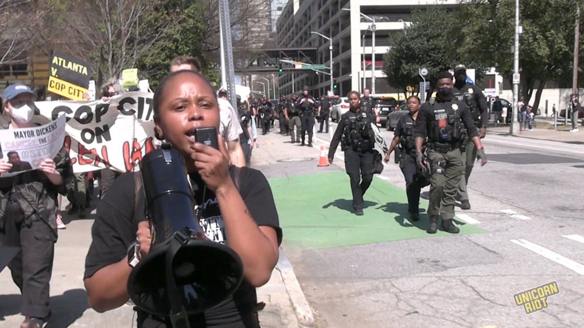 A small Stop Cop City protest march proceeds down an Atlanta sidewalk as several dozen police walk in the street to flank it. Some but not all officers have reinforced helmets and/or assault rifles. Most have plastic mass arrest ziptie handcuffs attached to their belt and/or vest. In this photo, Jasmine Burnett, a young black woman holding a megaphone, is leading the crowd. Burnett is Organizing Director of Community Movement Builders, an Atlanta-based "collective of black people creating sustainable, self-determining communities through cooperative economic advancement and collective community organizing," according to their website. 