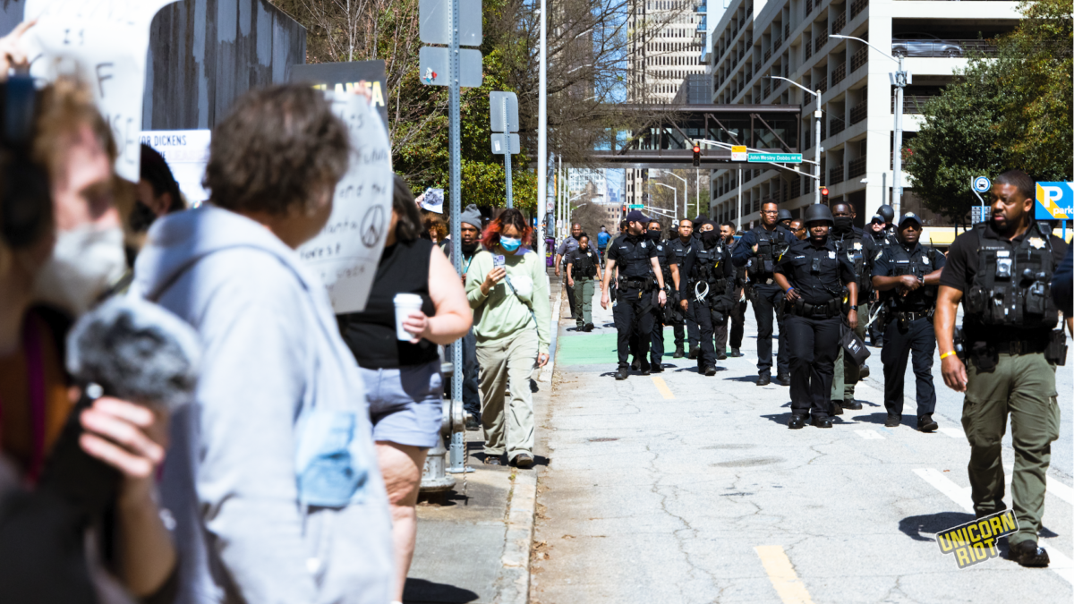 A small Stop Cop City protest march proceeds down an Atlanta sidewalk on March 7, 2023 as several dozen police walk in the street to flank it. Some but not all officers have reinforced helmets and/or assault rifles. Most have plastic mass arrest ziptie handcuffs attached to their belt and/or vest.