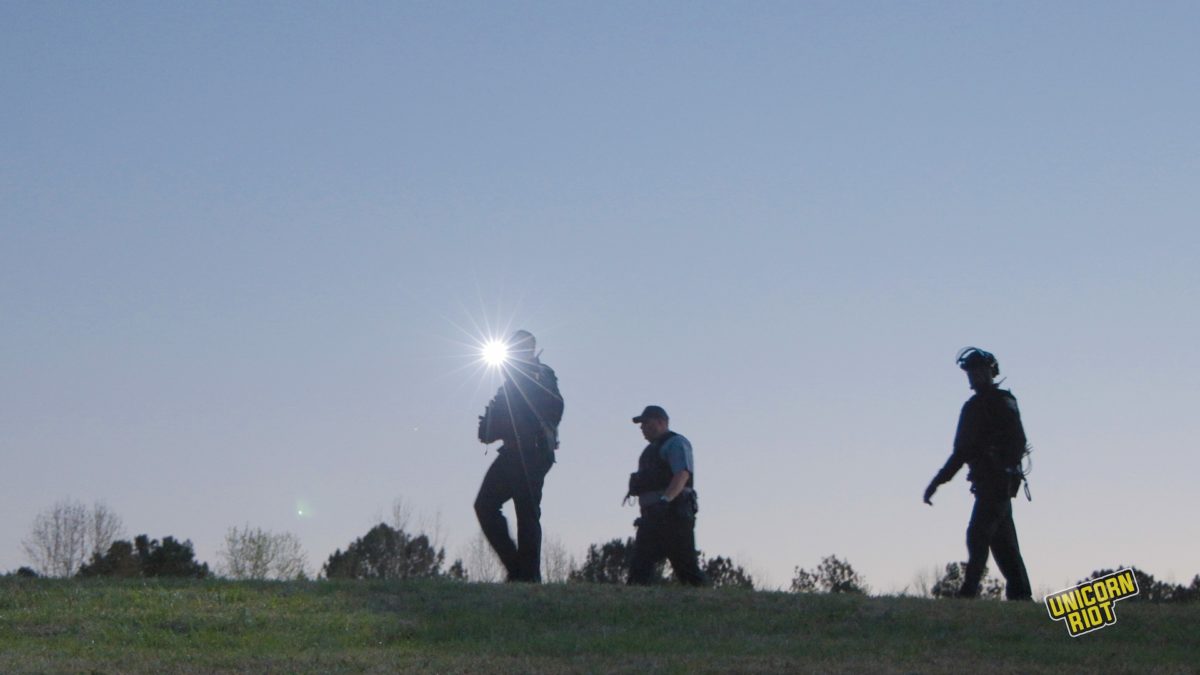 A photo taken from a lower vantage point shows three Georgia state troopers walking in the 'RC Field' clearing in Atlanta's South River Forest. Their darker outlines stand out against the blue evening sky. One of them, furthest to the right, has a riot helmet on; all 3 have plastic ziptie cuffs attached to their belts and/or vests. The leftmost officer is shining a flashlight towards the camera.