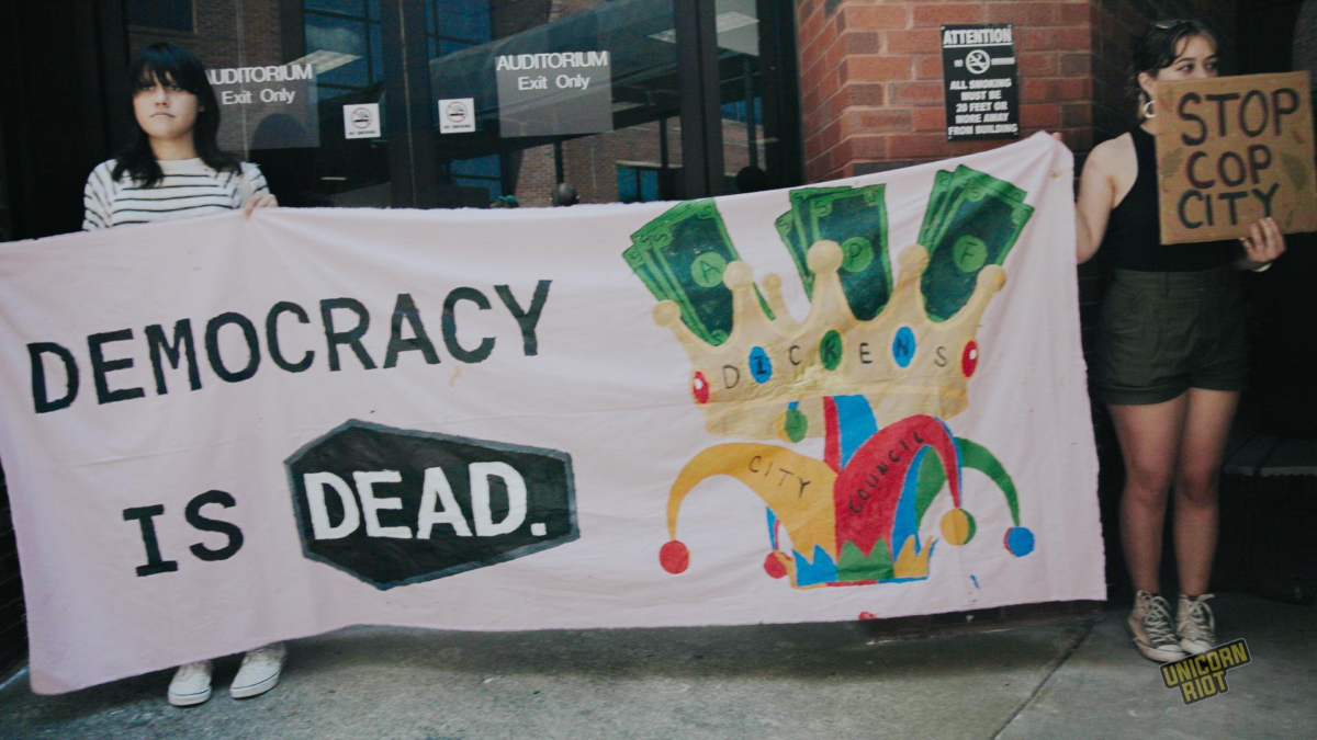 Protesters outside the DeKalb County Commissioners hold a banner made from white cloth with black text reading "Democracy is Dead" with "Dead" written in white against the black background of the shape of a coffin. To the right of the text is a jester's cap that's fused with a kings crown with stacks of cash sticking out from the top of the crown. Each of the multicolored jewels in the crown contains a letter - the crown spells out 'D-I-C-K-E-N-S' - a reference to Atlanta Mayor , 'Cop City' advocate and "sell-out" allegations denier Andre Dickens.