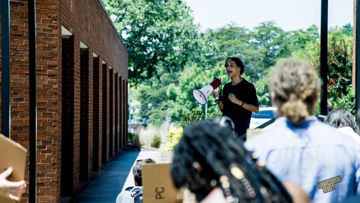 A young person with dark hair and a black shirt holds up a megaphone to address the crowd protesting outside the DeKalb County Commissioner's meeting at the county offices building.